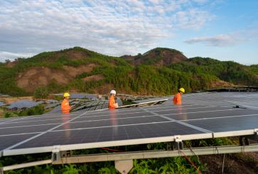 Three workers stand in between solar panels on top of a mountain.