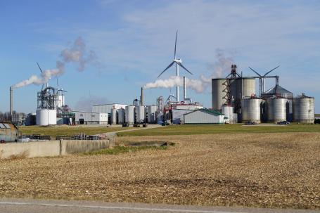 Smoke billows from an industrial plant producing ethanol fuel with corn fields in the foreground.