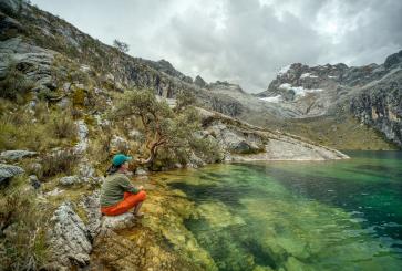 A person wearing a hat sits on the rocks overlooking Laguna Churup in Peru.