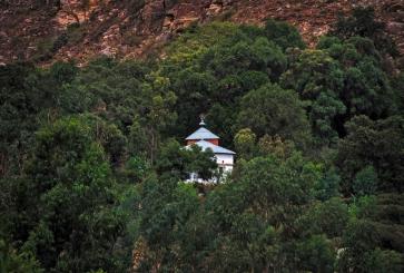 A small white church with a cross on top is perched on a steep mountainside encircled by trees. 